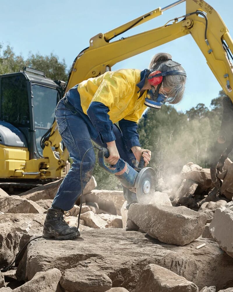 Lady sawing rock with safety equipment on, and digger machine in background.